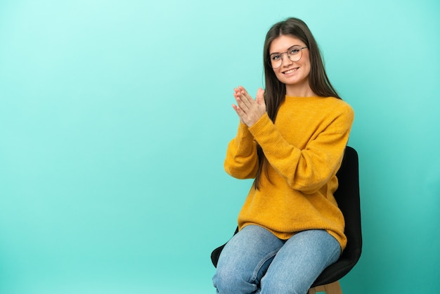 Young caucasian woman sitting on a chair isolated on blue wall applauding after presentation in a conference