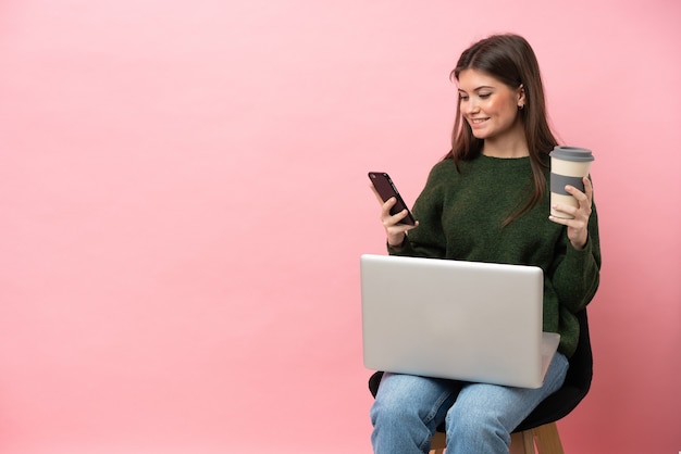 Young caucasian woman sitting on a chair with her laptop isolated on pink background holding coffee to take away and a mobile