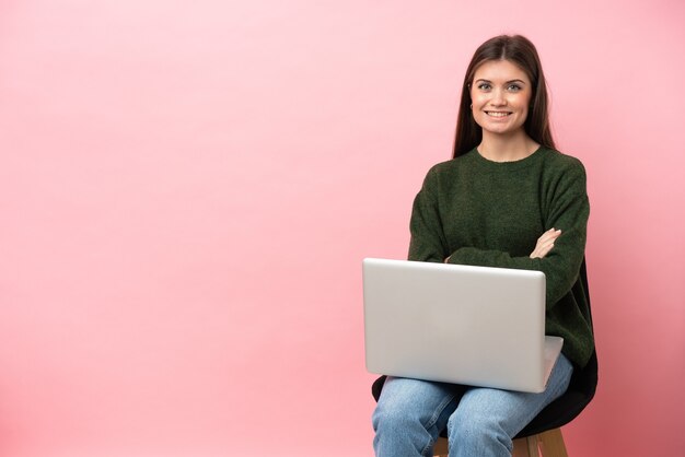 Young caucasian woman sitting on a chair with her laptop isolated on pink background keeping the arms crossed in frontal position