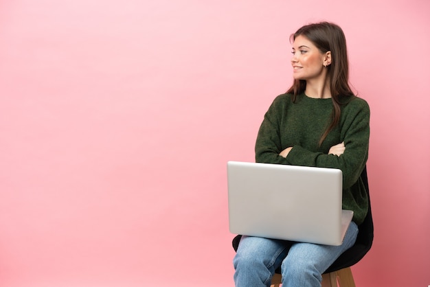 Young caucasian woman sitting on a chair with her laptop isolated on pink background in lateral position