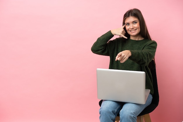 Young caucasian woman sitting on a chair with her laptop isolated on pink background making phone gesture and pointing front