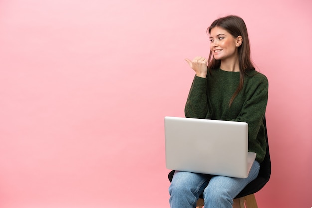 Young caucasian woman sitting on a chair with her laptop isolated on pink background pointing to the side to present a product