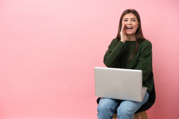 Giovane donna caucasica seduta su una sedia con il suo computer portatile isolato su sfondo rosa che grida con la bocca spalancata