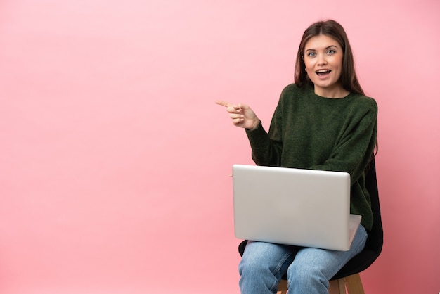 Young caucasian woman sitting on a chair with her laptop isolated on pink background surprised and pointing side