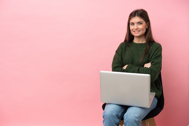 Young caucasian woman sitting on a chair with her laptop isolated on pink background with arms crossed and looking forward