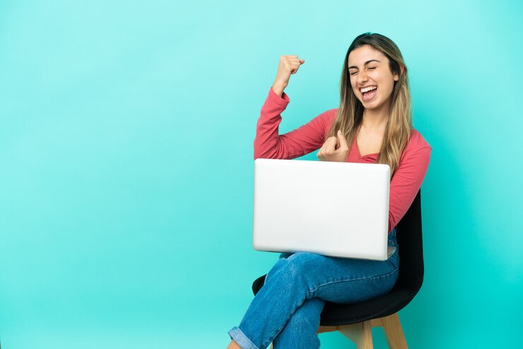 फोटो young caucasian woman sitting on a chair with her pc isolated on blue background celebrating a victory