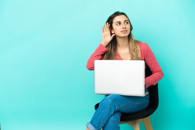 Young caucasian woman sitting on a chair with her pc isolated on blue background listening to something by putting hand on the ear