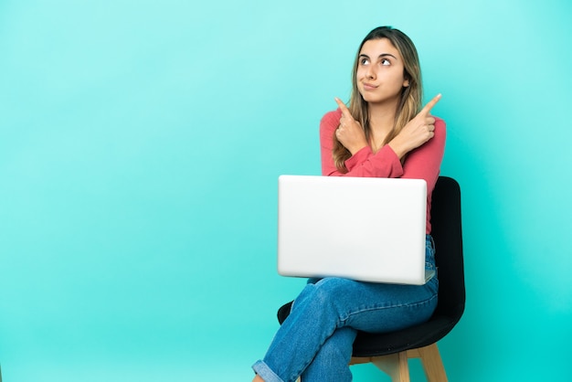 Young caucasian woman sitting on a chair with her pc isolated on blue background pointing to the laterals having doubts