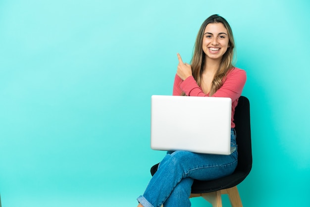 Young caucasian woman sitting on a chair with her pc isolated on blue background pointing to the side to present a product