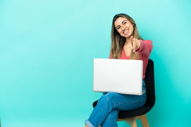 Young caucasian woman sitting on a chair with her pc isolated on blue background points finger at you with a confident expression