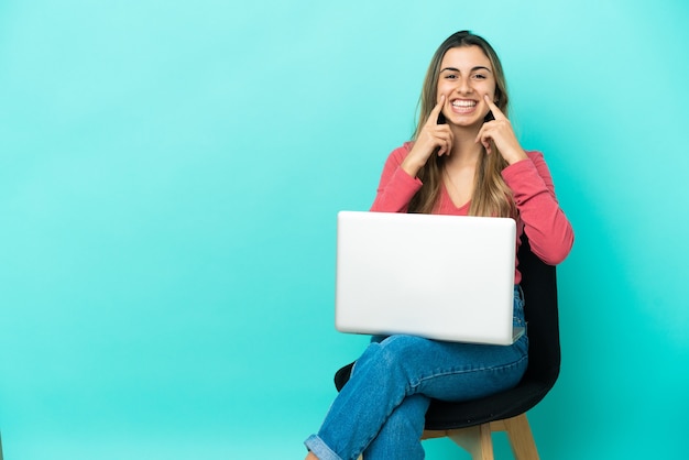 Молодая кавказская женщина сидит на стуле со своим компьютером, изолированным на синем фоне, улыбается со счастливым и приятным выражением лица