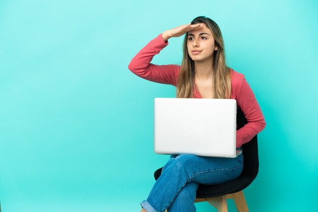 Young caucasian woman sitting on a chair with her pc isolated on blue wall looking far away with hand to look something