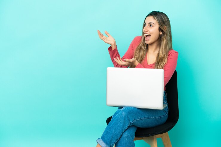 फोटो young caucasian woman sitting on a chair with her pc isolated on blue wall with surprise facial expression