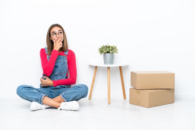 Young caucasian woman sitting on the floor among boxes having doubts and with confuse face expression