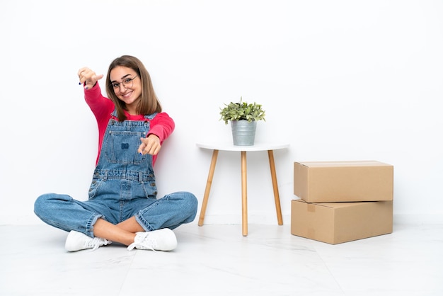 Young caucasian woman sitting on the floor among boxes points finger at you while smiling