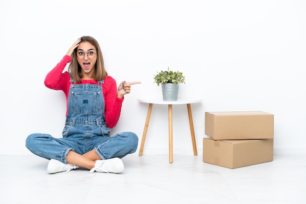 Young caucasian woman sitting on the floor among boxes surprised and pointing finger to the side