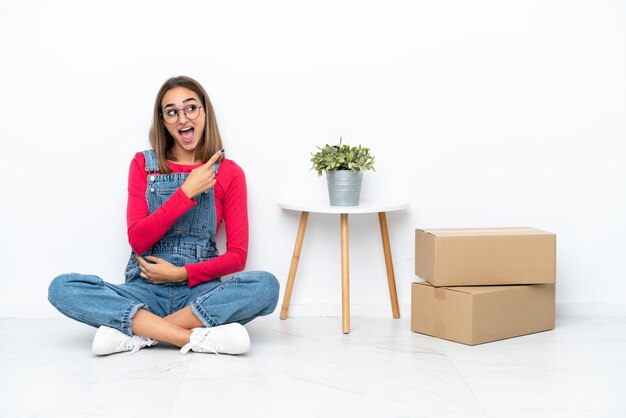 Young caucasian woman sitting on the floor among boxes surprised and pointing side