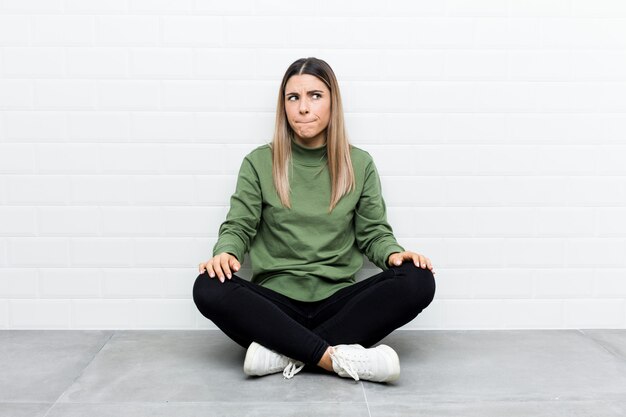 Young caucasian woman sitting on the floor confused, feels doubtful and unsure.