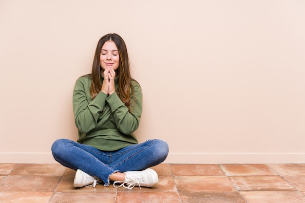 Young caucasian woman sitting on the floor holding hands in pray near mouth, feels confident.