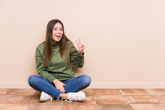 Young caucasian woman sitting on the floor isolated showing victory sign and smiling broadly.