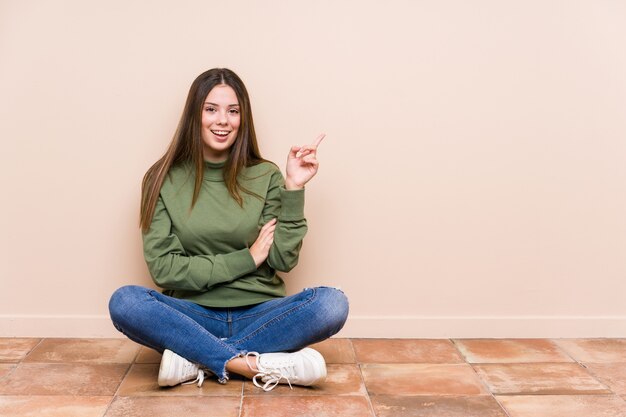 Young caucasian woman sitting on the floor isolated smiling cheerfully pointing with forefinger away.
