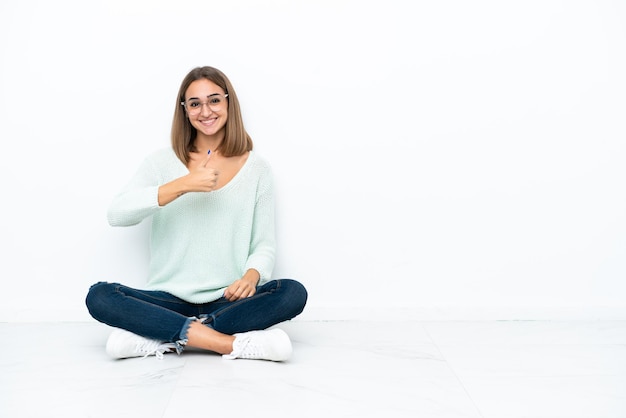 Young caucasian woman sitting on the floor isolated on white background giving a thumbs up gesture