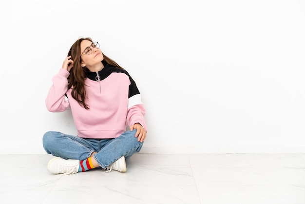 Young caucasian woman sitting on the floor isolated on white background having doubts and with confuse face expression