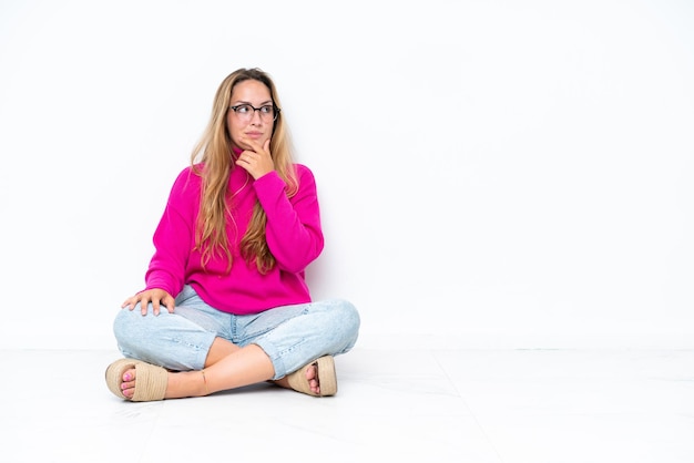 Young caucasian woman sitting on the floor isolated on white background having doubts and with confuse face expression