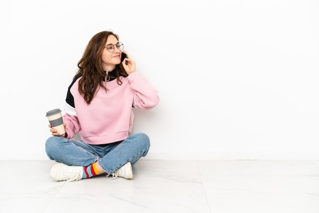 Young caucasian woman sitting on the floor isolated on white background holding coffee to take away and a mobile