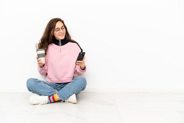 持ち帰り用のコーヒーと携帯電話を保持している白い背景で隔離の床に座っている若い白人女性