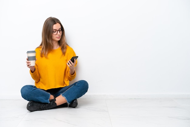 Young caucasian woman sitting on the floor isolated on white background holding coffee to take away and a mobile