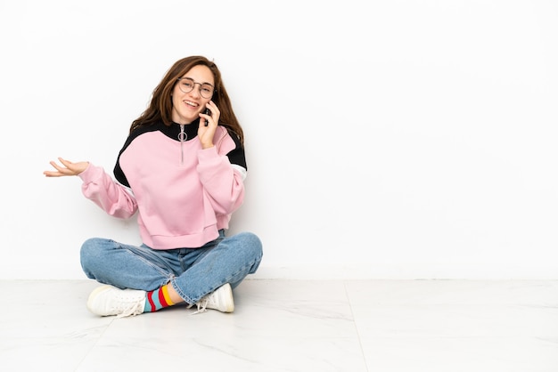 Молодая кавказская женщина сидит на полу, изолированном на белом фоне, разговаривает с кем-то по мобильному телефону