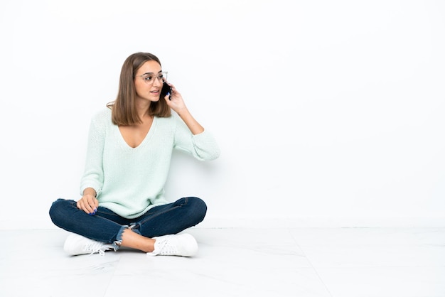 Young caucasian woman sitting on the floor isolated on white background keeping a conversation with the mobile phone
