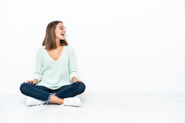 Young caucasian woman sitting on the floor isolated on white background laughing in lateral position