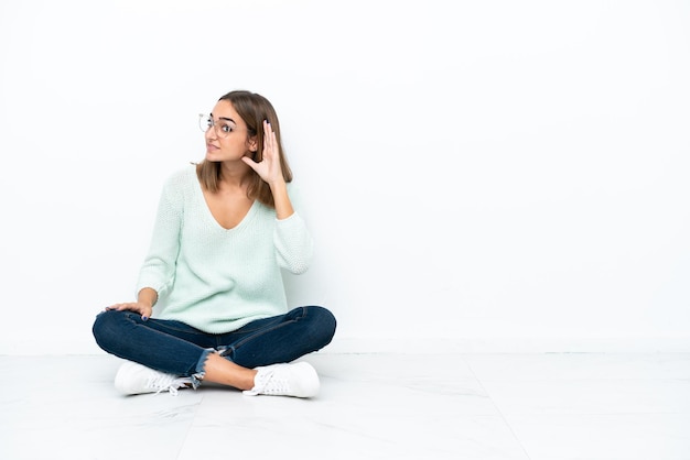 Foto giovane donna caucasica seduta sul pavimento isolato su sfondo bianco ascoltando qualcosa mettendo la mano sull'orecchio