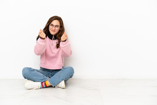 Young caucasian woman sitting on the floor isolated on white background making money gesture