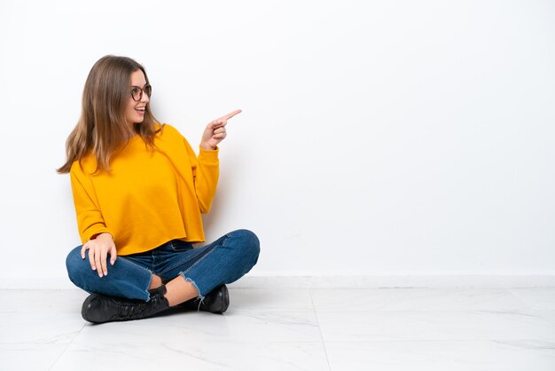 Young caucasian woman sitting on the floor isolated on white background pointing finger to the side and presenting a product