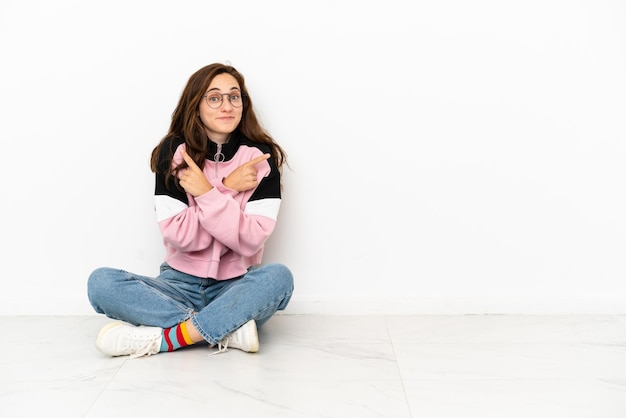 Foto giovane donna caucasica seduta sul pavimento isolato su sfondo bianco che punta ai laterali che hanno dubbi