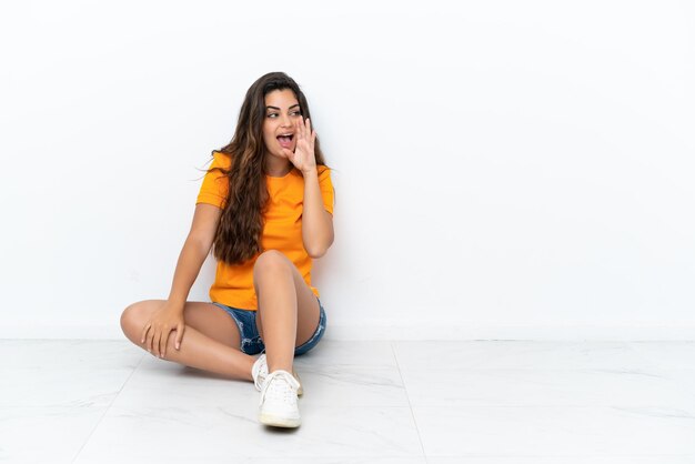 Young caucasian woman sitting on the floor isolated on white background shouting with mouth wide open to the lateral