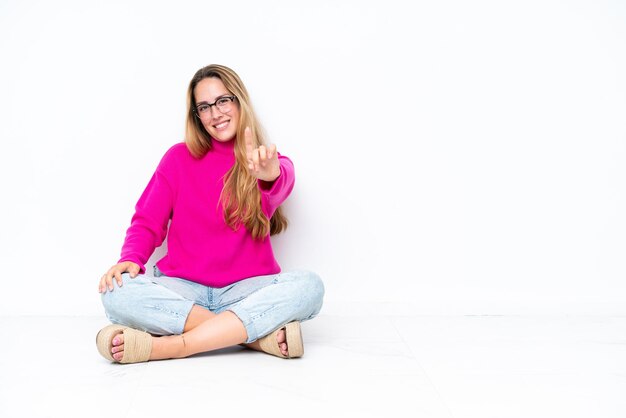 Foto giovane donna caucasica seduta sul pavimento isolato su sfondo bianco che mostra e solleva un dito