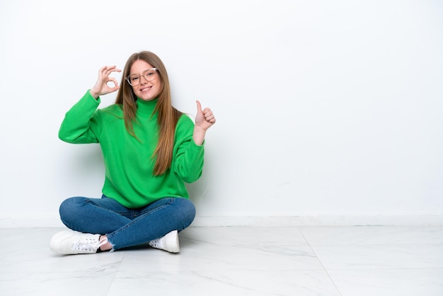 Young caucasian woman sitting on the floor isolated on white background showing ok sign and thumb up gesture