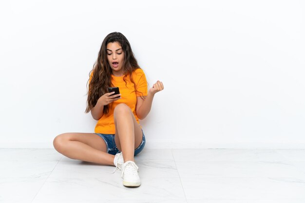 Young caucasian woman sitting on the floor isolated on white background surprised and sending a message