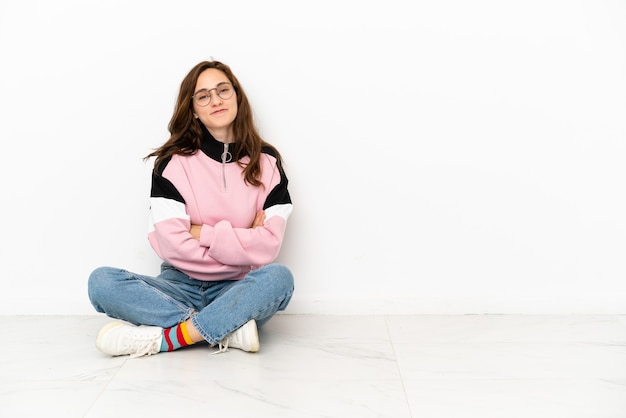 Young caucasian woman sitting on the floor isolated on white background with arms crossed and looking forward