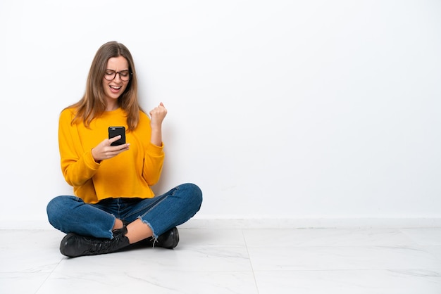 Young caucasian woman sitting on the floor isolated on white background with phone in victory position