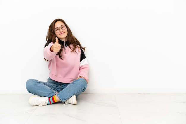 Photo young caucasian woman sitting on the floor isolated on white background with thumbs up because something good has happened