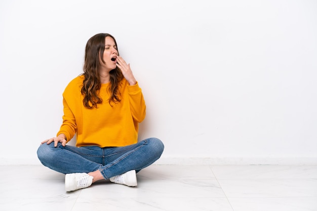 Young caucasian woman sitting on the floor isolated on white wall yawning and covering wide open mouth with hand