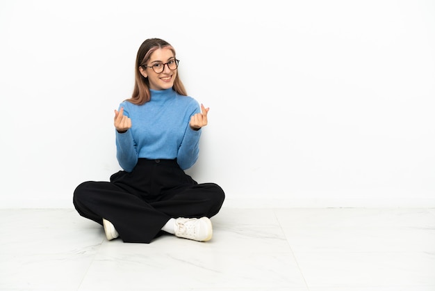 Young Caucasian woman sitting on the floor making money gesture