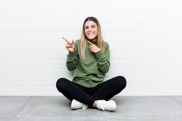 Young caucasian woman sitting on the floor pointing with forefingers to a copy space, expressing excitement and desire.