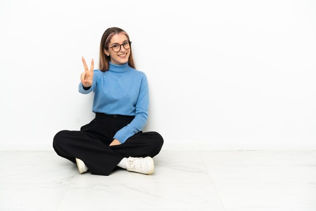 Young Caucasian woman sitting on the floor smiling and showing victory sign