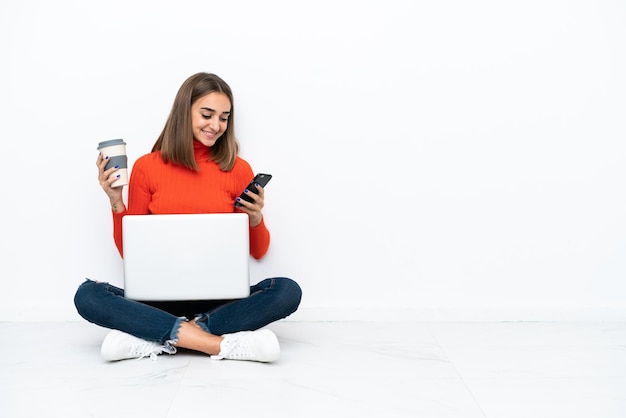 Young caucasian woman sitting on the floor with a laptop holding coffee to take away and a mobile